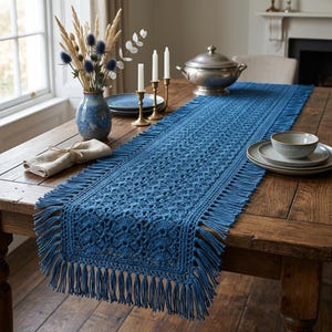 May include: A rectangular blue crochet table runner featuring an intricate openwork pattern and long fringe along all edges. The runner is laid out flat on a wooden table, showcasing the detailed yarn texture and the consistent deep blue color.