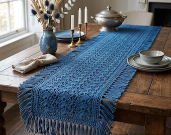 May include: A rectangular blue crochet table runner featuring an intricate openwork pattern and long fringe along all edges. The runner is laid out flat on a wooden table, showcasing the detailed yarn texture and the consistent deep blue color.
