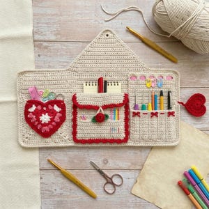 May include: A beige crochet organizer case featuring a heart-shaped pocket, a central flap pocket with a red rose accent, and a side section for tools. The case includes a red heart button closure, a gold crochet hook, and metal scissors.