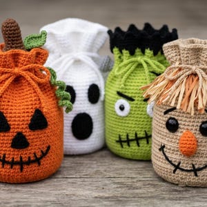 May include: Four crochet drawstring bags featuring Halloween characters: an orange pumpkin with a brown stem, a white ghost with black eyes and mouth, a green monster with black hair and stitches, and a tan scarecrow with a raffia fringe.