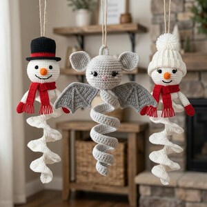 May include: Three hanging crochet ornaments: a snowman wearing a black top hat and red scarf, a gray bat with large ears and wings, and a snowman wearing a white pom-pom beanie and red scarf. Each figure features a white spiral tail hanging downward.