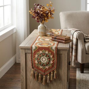 Puede incluir: Un camino de mesa de ganchillo rectangular con un patrón de mandala repetitivo en tonos óxido, verde salvia, crema y amarillo mostaza. El camino de mesa está acabado con un borde puntiagudo y borlas de hilo decorativas colgando.