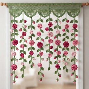 May include: A crochet window valance featuring a scalloped green top edge with hanging vertical strands of green vines, leaves, and various shades of pink and red roses. The piece is suspended from a wooden rod across a white door frame.