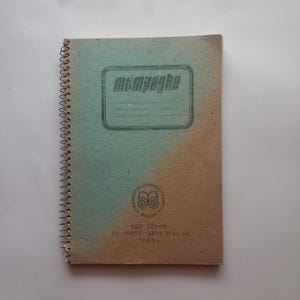 Può includere: Un quaderno a spirale con una copertina bicolore caratterizzata da una sfumatura dal blu chiaro al beige. La copertina mostra il testo cirillico "тетрадка" all'interno di una cornice rettangolare, un logo circolare e testo "1986 г."