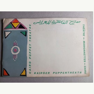 May include: A rectangular paper program featuring a blue vertical panel on the left with colorful geometric shapes and a central circular emblem. The text reads Cairo Puppet Theatre, Théâtre des Marionnettes du Caire, Kairoer Puppentheater, and Arabic script.