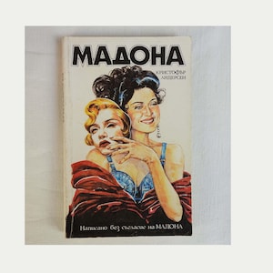May include: A paperback book titled MADONA by Christopher Andersen featuring a cover illustration of two figures, one with blonde hair and one with dark hair, wearing a blue top and a dark red wrap, with Cyrillic text printed on the front cover.
