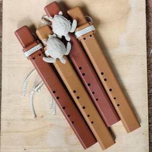 May include: Four brown flutes with a turtle charm and a white rope detail. The flutes are in varying shades of brown, with a series of holes along the body. The turtle charms are light gray. The flutes are arranged diagonally on a wooden surface.