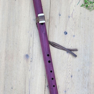 May include: A purple wooden flute with six finger holes and a leather strap. The flute is lying on a wooden surface.