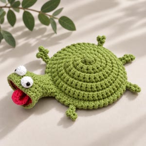 May include: A green crocheted turtle-shaped coaster featuring a spiral-patterned shell, four small legs, and a head with two white protruding eyes with black pupils and an open mouth displaying a bright pink tongue, crafted from textured yarn.