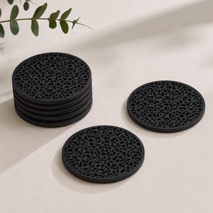 May include: A set of circular black coasters featuring a repeating intricate floral cutout pattern. The collection includes a stack of five coasters and two individual coasters laid out flat, all displaying the same detailed openwork design.