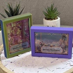 May include: Two rectangular framed art pieces. The left frame is green and displays an illustration titled Witch's Tea House, measuring 25.4 cm x 35.6 cm. The right frame is purple and displays an illustration titled Dreamy Workspace, measuring 25.4 cm x 35.6 cm.