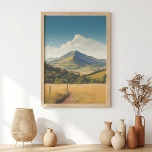 May include: A framed landscape art print featuring a mountain peak under a blue sky with white clouds, overlooking a golden grassy field, a dirt path, and green trees. The artwork is displayed in a light wood frame on a white wall.