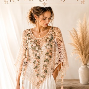 Può includere: Uno scialle all'uncinetto beige intitolato Rosabelle Bloom Shawl, caratterizzato da un motivo a rete a maglia aperta decorato con delicati tralci floreali ricamati in tonalità rosa tenue e verde, rifinito con piccole nappe pendenti sui bordi.