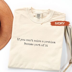 Può includere: Una t-shirt a maniche corte color avorio con scollo rotondo e un'etichetta del marchio Comfort Colors sul colletto interno. La parte anteriore presenta un testo nero in stile macchina da scrivere: If you can't solve a problem, Become part of it.