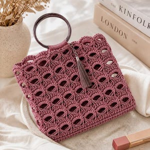 May include: A dusty rose handbag crafted with an intricate openwork crochet pattern. The bag features a circular dark metallic handle and a matching tassel charm attached to the top. The square-shaped design showcases a repeating knotted texture.