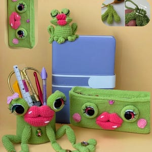 May include: A crochet FROGY SET featuring a green frog-shaped pencil holder with large eyes, pink lips, and a small pink bow, alongside a matching rectangular green zippered pouch with similar facial features and a small frog figurine.