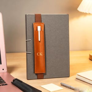 May include: A gray notebook with a vertical brown leather pen holder strap featuring the gold embossed initials CK. The strap is secured with an elastic band and holds a white pen. The notebook has a textured cover and a simple, functional design.