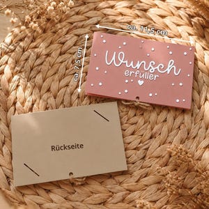 Peut inclure: Une carte rectangulaire rose poudr&eacute; avec le texte blanc "Wunsch erf&uuml;ller", un petit c&oelig;ur et des points blancs. Elle mesure 11,5 cm sur 7,5 cm. En dessous se trouve une carte beige clair marqu&eacute;e "R&uuml;ckseite" avec des fentes aux coins.