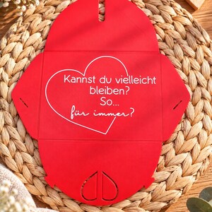 May include: A bright red, unfolded paper envelope with a heart-shaped cutout at the bottom. The center features a white heart outline containing the German text: "Kannst du vielleicht bleiben? So... f&uuml;r immer?"