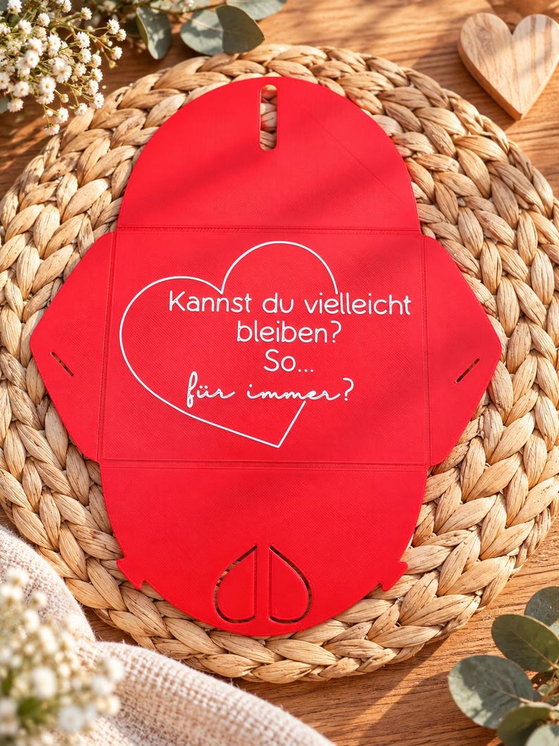 May include: A bright red, unfolded paper envelope with a heart-shaped cutout at the bottom. The center features a white heart outline containing the German text: "Kannst du vielleicht bleiben? So... f&uuml;r immer?"
