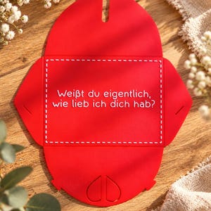 May include: A flat, red, die-cut cardstock envelope template featuring a white dashed rectangular border and the German text "Wei&szlig;t du eigentlich, wie lieb ich dich hab?" printed in white. The bottom flap displays two embossed heart shapes.