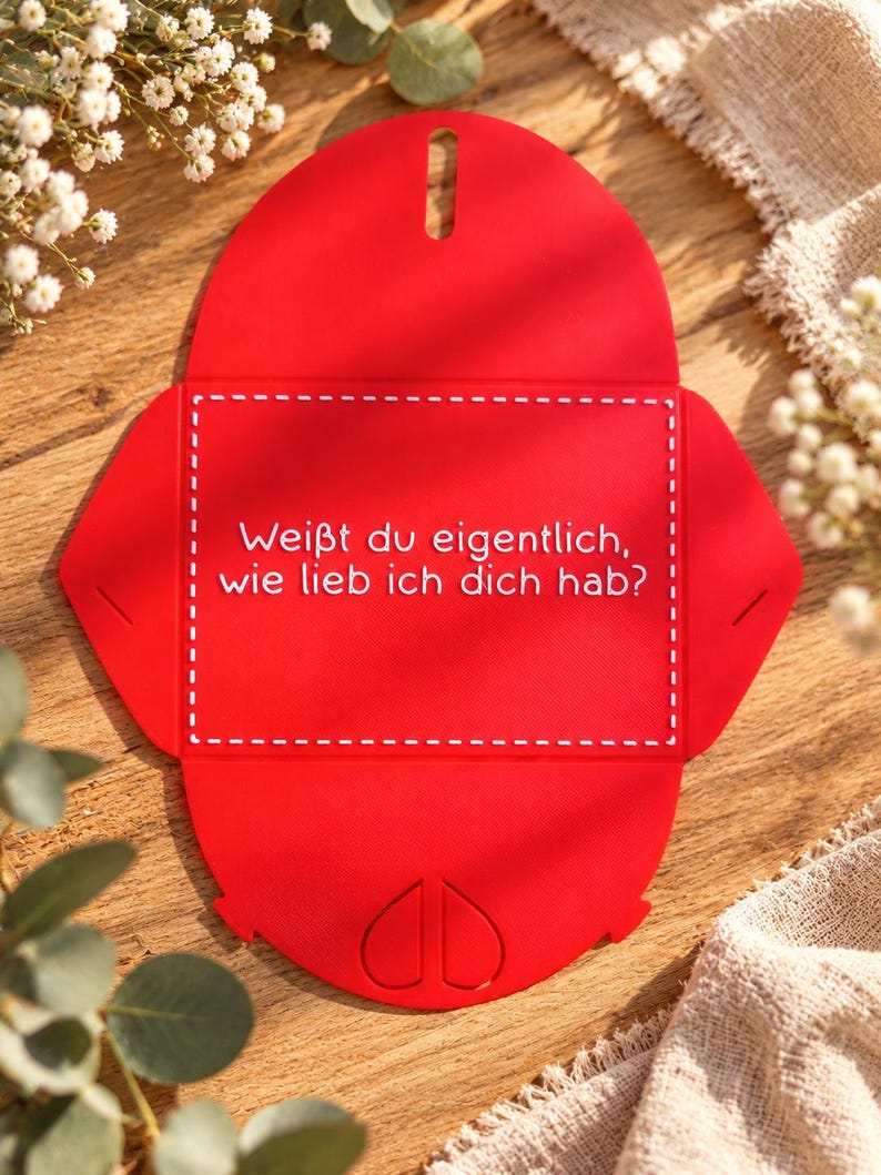 May include: A flat, red, die-cut cardstock envelope template featuring a white dashed rectangular border and the German text "Wei&szlig;t du eigentlich, wie lieb ich dich hab?" printed in white. The bottom flap displays two embossed heart shapes.