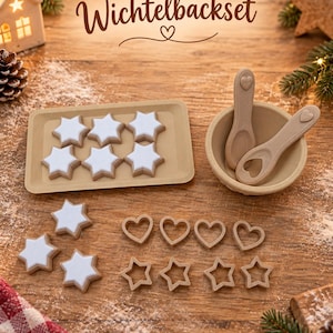 May include: A baking set labeled Wichtelbackset featuring a rectangular tray with six white star-shaped cookies, a small bowl with two spoons, four heart-shaped cookie cutters, and four star-shaped cookie cutters on a wooden surface.