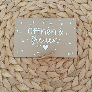 Peut inclure: Un panneau rectangulaire beige avec le texte blanc "&Ouml;ffnen & freuen" et une petite ic&ocirc;ne de c&oelig;ur blanc sous le texte. Le panneau est d&eacute;cor&eacute; de points blancs dispers&eacute;s et comporte un petit n&oelig;ud en ficelle de jute au centre en bas.