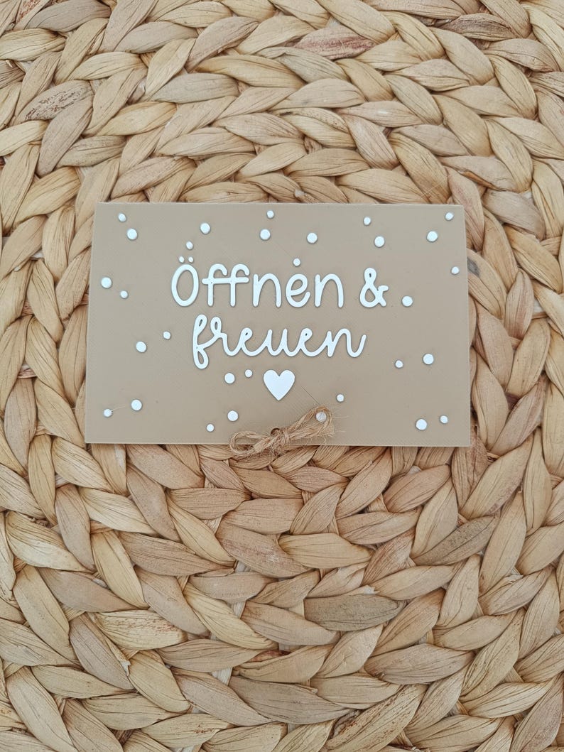 Peut inclure: Un panneau rectangulaire beige avec le texte blanc "&Ouml;ffnen & freuen" et une petite ic&ocirc;ne de c&oelig;ur blanc sous le texte. Le panneau est d&eacute;cor&eacute; de points blancs dispers&eacute;s et comporte un petit n&oelig;ud en ficelle de jute au centre en bas.