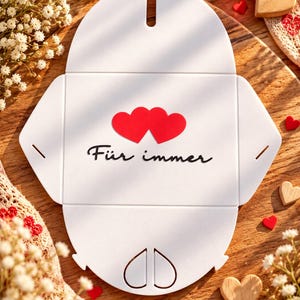 May include: A white, flat-packed cardboard gift box with a die-cut oval shape. The center features two red hearts and the black text "F&uuml;r immer". The box has pre-scored folding lines and a small slot at the top for assembly.