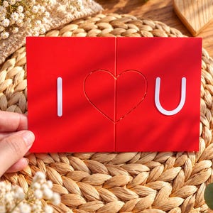May include: A rectangular red greeting card featuring the white text I and U separated by a central heart shape. The card is divided into two vertical panels that meet in the middle, forming a complete heart graphic when placed together.