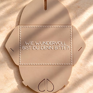 May include: A beige, flat, unfolded envelope-shaped object made of a smooth material. It features a central rectangular area with a dashed border and the German text "WIE WUNDERVOLL BIST DU DENN BITTE?!" printed in white, with slots for folding.