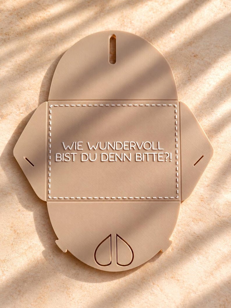 May include: A beige, flat, unfolded envelope-shaped object made of a smooth material. It features a central rectangular area with a dashed border and the German text "WIE WUNDERVOLL BIST DU DENN BITTE?!" printed in white, with slots for folding.