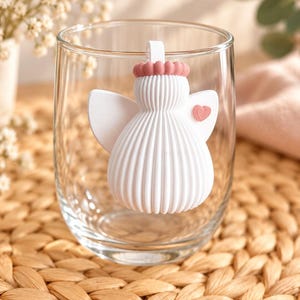May include: A clear glass tumbler featuring a white, ribbed, angel-shaped silicone attachment. The angel has a small pink halo, a pink heart detail on its wing, and a white loop at the top for hanging or securing to the rim of the glass.