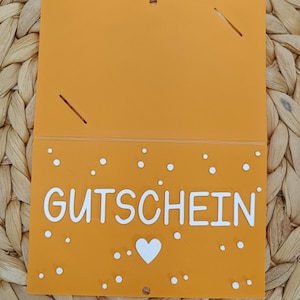 Peut inclure: Une carte rectangulaire orange avec le mot GUTSCHEIN imprim&eacute; en lettres capitales blanches. La carte pr&eacute;sente une ic&ocirc;ne de c&oelig;ur blanc sous le texte et de petits points blancs dispers&eacute;s sur la moiti&eacute; inf&eacute;rieure. Il y a deux fentes et un trou.