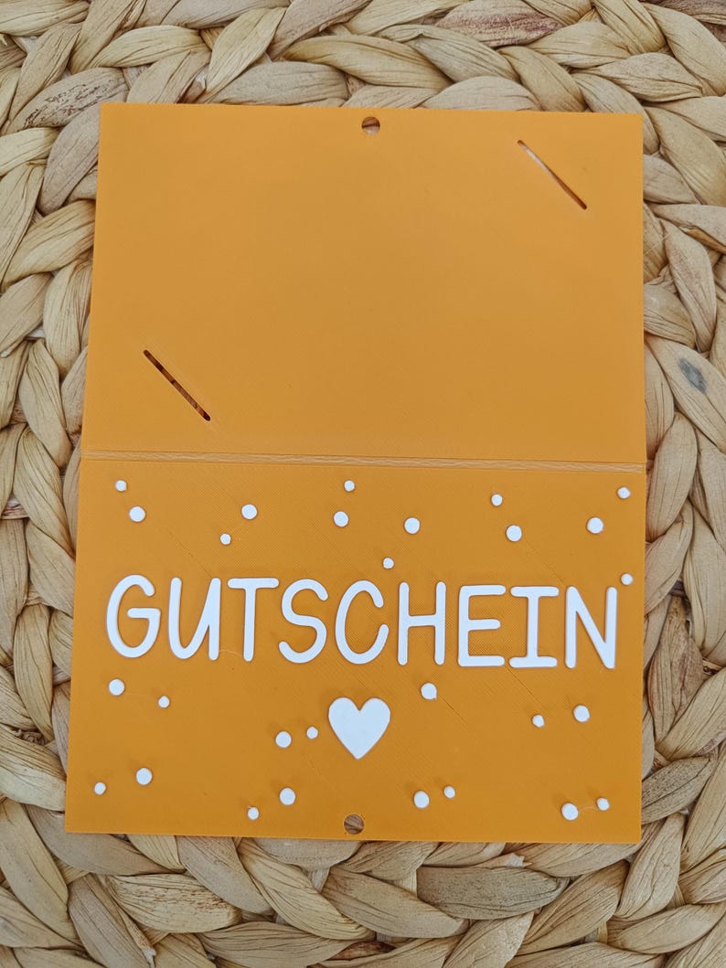 Peut inclure: Une carte rectangulaire orange avec le mot GUTSCHEIN imprim&eacute; en lettres capitales blanches. La carte pr&eacute;sente une ic&ocirc;ne de c&oelig;ur blanc sous le texte et de petits points blancs dispers&eacute;s sur la moiti&eacute; inf&eacute;rieure. Il y a deux fentes et un trou.