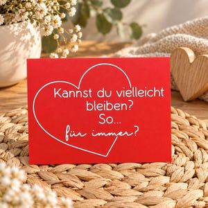 May include: A rectangular red greeting card featuring a white outline of a heart and the German text: Kannst du vielleicht bleiben? So... f&uuml;r immer? The card is positioned on a round woven straw mat.