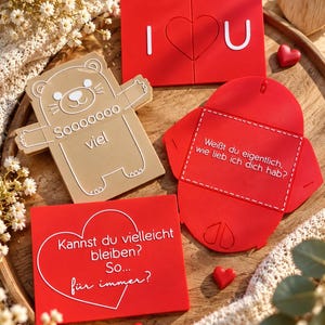 May include: A collection of flat, rectangular and shaped items in red and beige. One red piece reads I heart U, a beige bear-shaped piece reads Sooooooo viel, a red envelope reads Wei&szlig;t du eigentlich, wie lieb ich dich hab?, and a red card reads Kannst du vielleicht bleiben? So... f&uuml;r immer?
