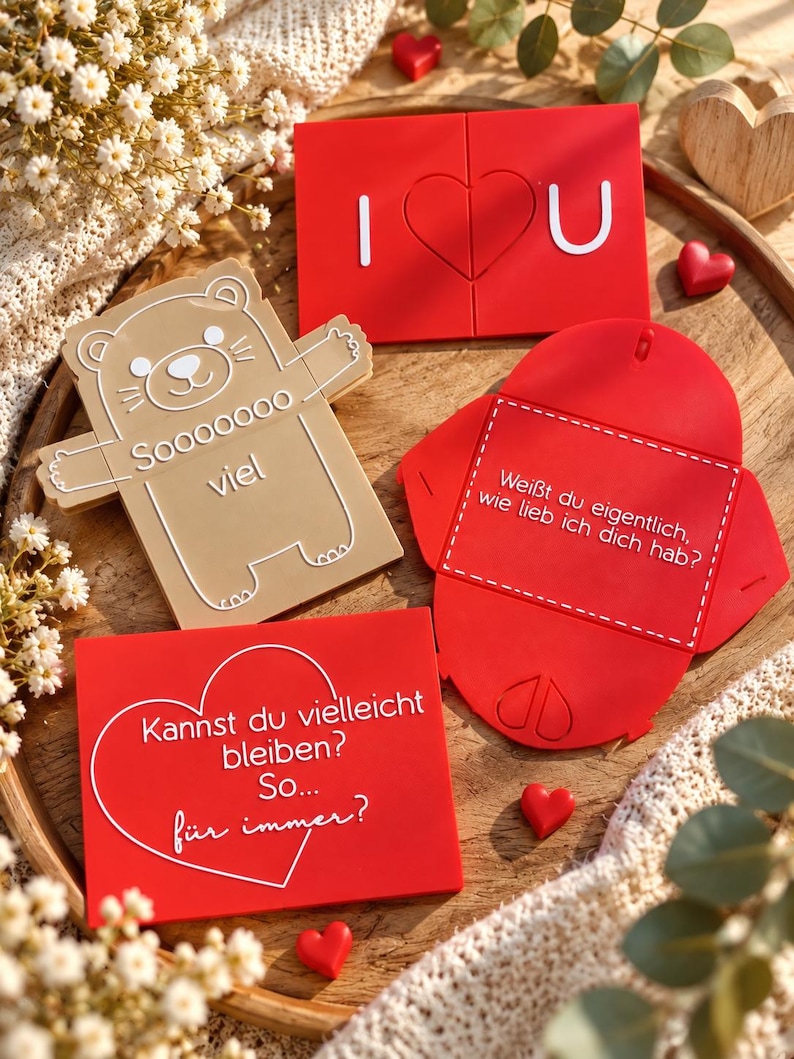 May include: A collection of flat, rectangular and shaped items in red and beige. One red piece reads I heart U, a beige bear-shaped piece reads Sooooooo viel, a red envelope reads Wei&szlig;t du eigentlich, wie lieb ich dich hab?, and a red card reads Kannst du vielleicht bleiben? So... f&uuml;r immer?