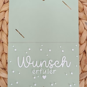 Peut inclure: Une carte rectangulaire vert sauge avec le texte blanc en relief "Wunsch erf&uuml;ller" au centre. La carte est d&eacute;cor&eacute;e de petits points blancs et d'un c&oelig;ur blanc. La partie sup&eacute;rieure comporte deux fentes diagonales et un petit trou de suspension.