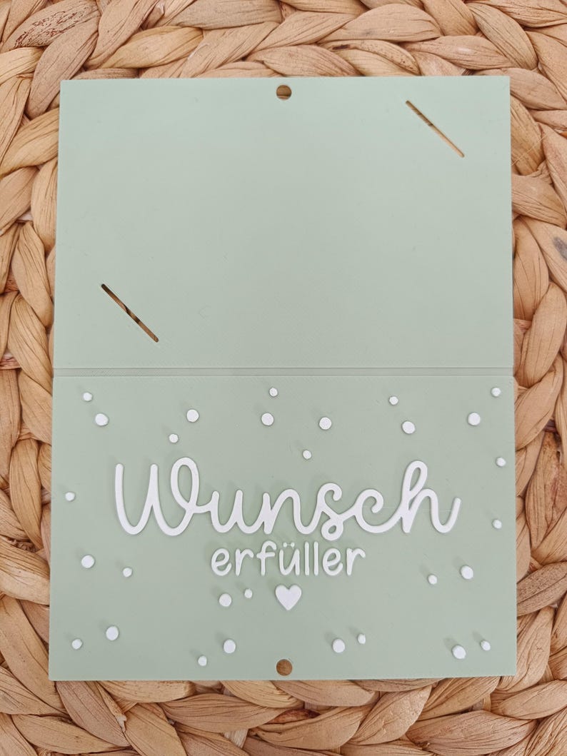Peut inclure: Une carte rectangulaire vert sauge avec le texte blanc en relief "Wunsch erf&uuml;ller" au centre. La carte est d&eacute;cor&eacute;e de petits points blancs et d'un c&oelig;ur blanc. La partie sup&eacute;rieure comporte deux fentes diagonales et un petit trou de suspension.