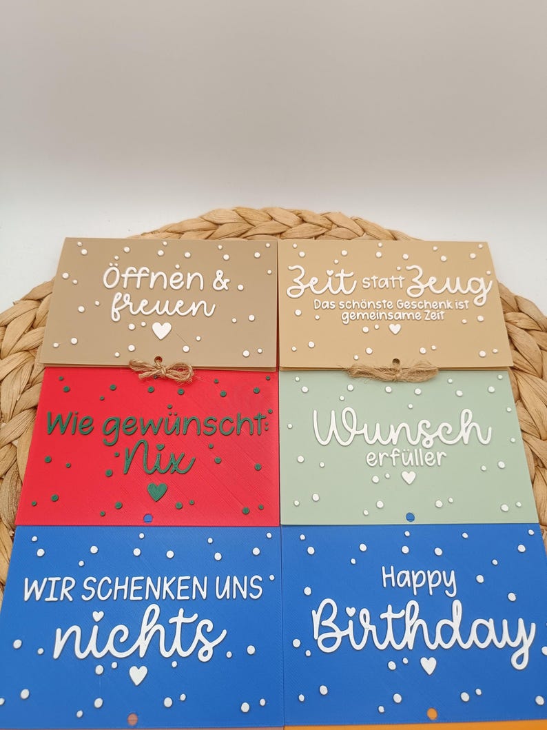 Peut inclure: Six cartes rectangulaires de couleur beige, rouge, vert clair et bleu, orn&eacute;es de textes en blanc ou vert et de petits c&oelig;urs. Les cartes portent les inscriptions "&Ouml;ffnen & freuen," "Zeit statt Zeug," "Wie gew&uuml;nscht: Nix," "Wunsch erf&uuml;ller," "WIR SCHENKEN UNS nichts" et "Happy Birthday."