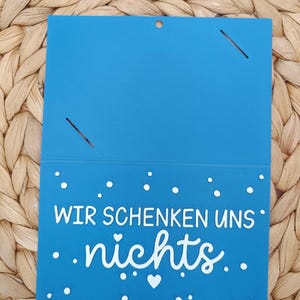Peut inclure: Une carte rectangulaire bleue avec un pli horizontal, pr&eacute;sentant le texte blanc "WIR SCHENKEN UNS nichts" ainsi que de petits points blancs et une ic&ocirc;ne de c&oelig;ur. La partie sup&eacute;rieure comporte deux fentes diagonales et un petit trou central.