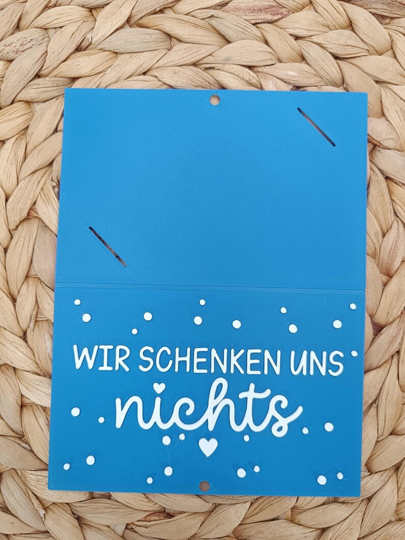 Peut inclure: Une carte rectangulaire bleue avec un pli horizontal, pr&eacute;sentant le texte blanc "WIR SCHENKEN UNS nichts" ainsi que de petits points blancs et une ic&ocirc;ne de c&oelig;ur. La partie sup&eacute;rieure comporte deux fentes diagonales et un petit trou central.