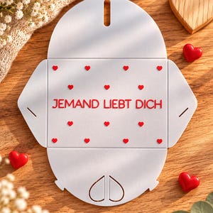 May include: A white, flat, die-cut paper envelope template featuring a repeating pattern of small red hearts and the red text JEMAND LIEBT DICH centered on the front panel. The design includes a heart-shaped cutout at the bottom flap.