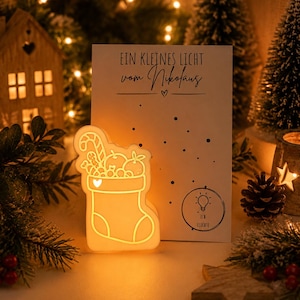 May include: A white, glowing, stocking-shaped LED light featuring an outline of a candy cane and fruit, positioned in front of a white card with the text "EIN KLEINES LICHT vom Nikolaus" and a small lightbulb icon with the text "ICH LEUCHTE".
