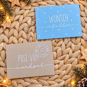 Könnte beinhalten: Eine hellblaue Karte mit weißen Sternen und dem Text WUNSCH erfüller sowie eine beige Karte mit dem Text POST VOM nordpol und einem runden Stempel mit der Aufschrift NORDPOL 2412 POSTAMT, beide mit weißer, erhabener Schrift versehen.