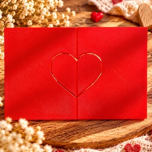 May include: A rectangular red greeting card featuring a centered heart outline formed by two halves that meet in the middle. The card has a textured surface and is positioned horizontally, displaying a clean, minimalist design with a vibrant red finish.