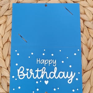 Peut inclure: Une carte rectangulaire bleue avec le texte blanc Happy Birthday &eacute;crit dans une police stylis&eacute;e. La partie inf&eacute;rieure est d&eacute;cor&eacute;e de petits points blancs et d'un c&oelig;ur. La partie sup&eacute;rieure comporte deux fentes diagonales et un petit trou.