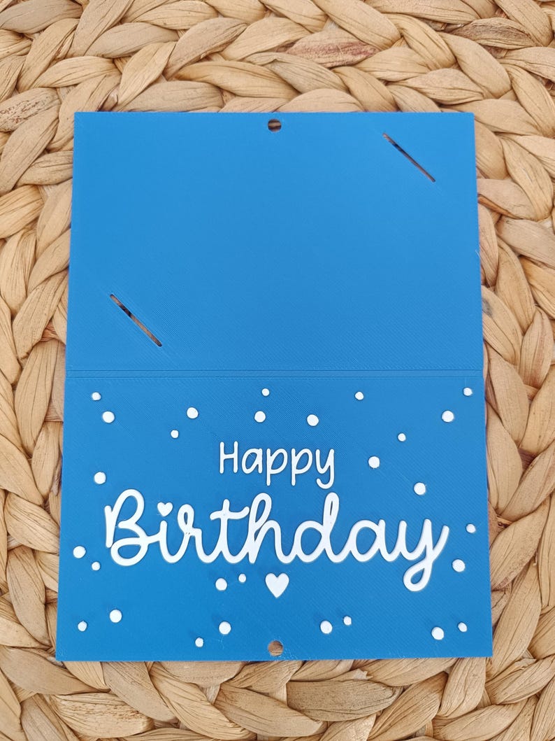 Peut inclure: Une carte rectangulaire bleue avec le texte blanc Happy Birthday &eacute;crit dans une police stylis&eacute;e. La partie inf&eacute;rieure est d&eacute;cor&eacute;e de petits points blancs et d'un c&oelig;ur. La partie sup&eacute;rieure comporte deux fentes diagonales et un petit trou.